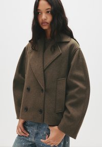 Brown, wool-blend double-breasted coat with a notched collar, large buttons, and a boxy fit. Features a side pocket and textured fabric.