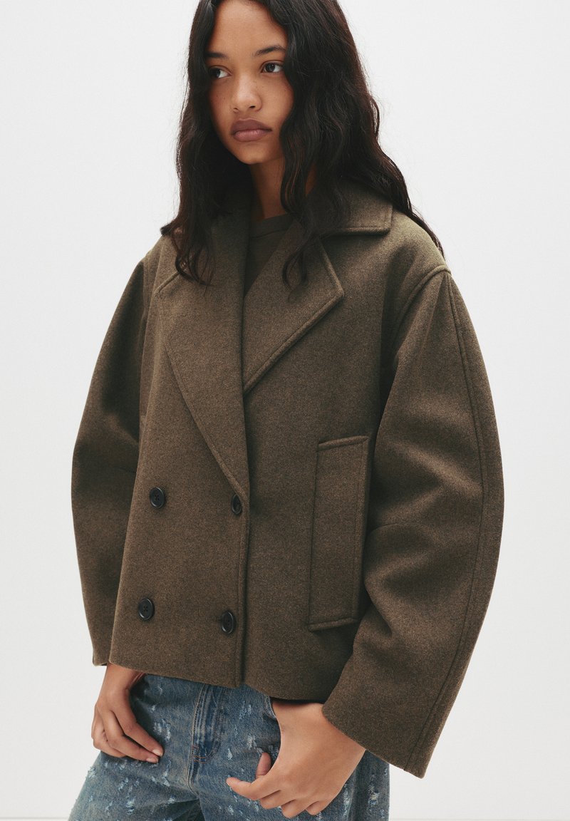 Brown, wool-blend double-breasted coat with a notched collar, large buttons, and a boxy fit. Features a side pocket and textured fabric.
