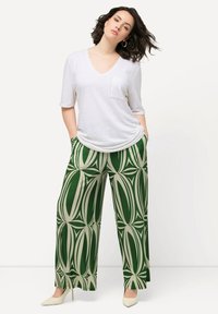 Weißes V-Ausschnitt-T-Shirt mit Tasche, kombiniert mit weiten grünen Hosen mit großen cremefarbenen geometrischen Mustern. Das Model trägt beigefarbene Absatzschuhe.