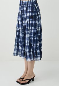 Gonna midi con motivo tie-dye bianco e blu navy, dettagli plissettati, indossata con sandali con tacco neri su uno sfondo chiaro.