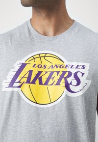 Szara bawełniana koszulka na ramiączkach z dużą żółtą grafiką koszykówki oraz napisem "LOS ANGELES LAKERS" w purpurowym kolorze. Miękka tekstura, casualowy design.