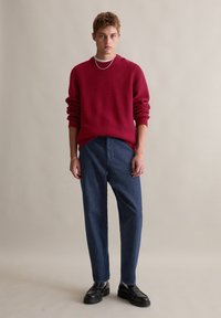 Roter strukturierten Strickpullover mit langen Ärmeln, kombiniert mit lockeren, dunkelblauen Jeans und schwarzen Schuhen. Minimalistische silberne Kettenhalskette.