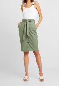Grüne knielange Schürze aus leichtem Stoff mit einem gebundenen Taillenband, Knopfdetails vorne und seitlichen Taschen. Tragen mit einem weißen Tanktop.