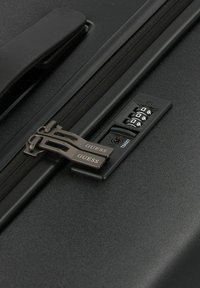 Cerniera della valigia nera con due tiranti metallici che presentano il logo "GUESS", una combinazione di lucchetto con tre dischi e superficie esterna texture.