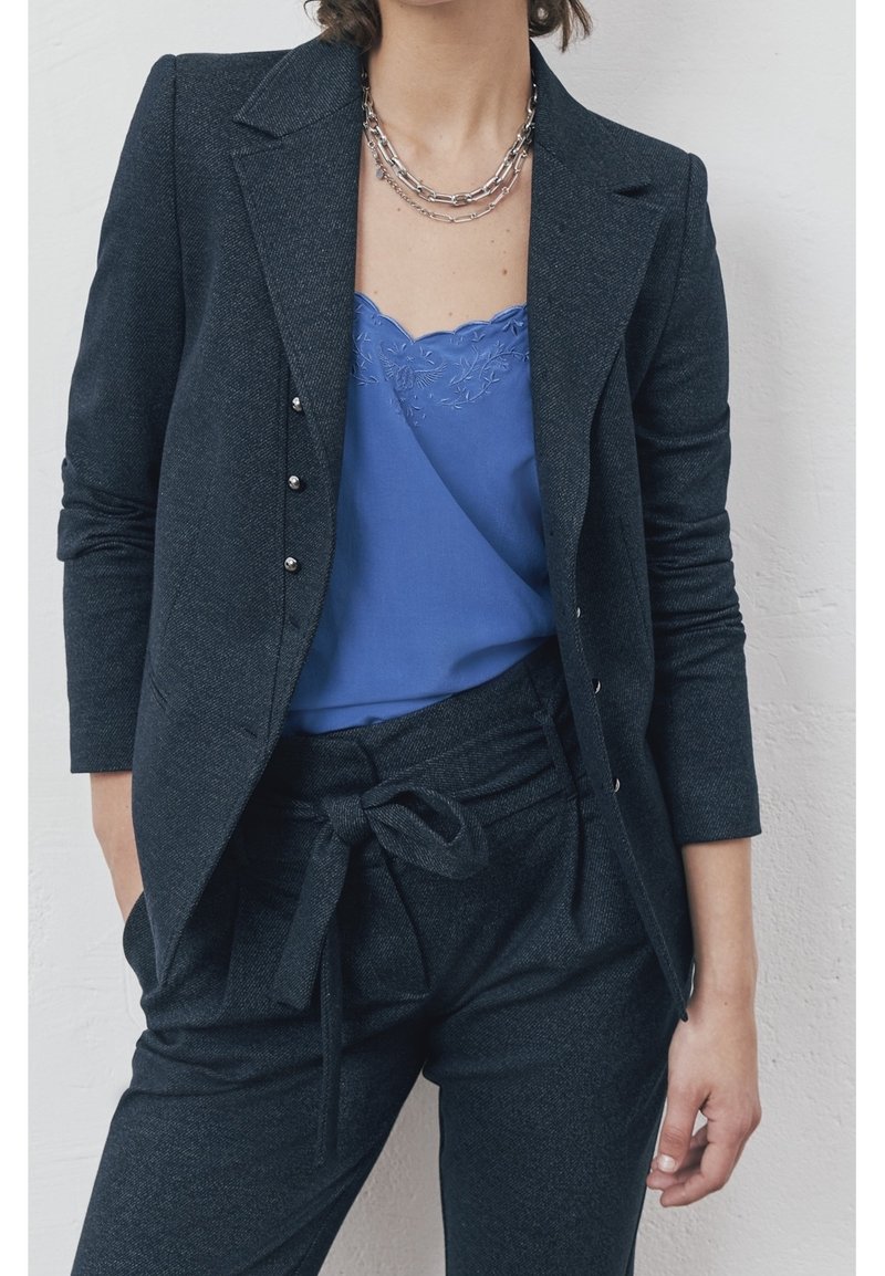 Femme portant un costume gris foncé avec un haut bleu brodé orné de dentelle et deux colliers en chaîne argentés, debout, une main dans la poche.