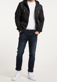 Mężczyzna w czarnej kurtce zapinanej na zamek, białej koszulce, ciemnoniebieskich, podwiniętych dżinsach, białych skarpetkach i białych sneakersach stoi z rękami w kieszeniach.