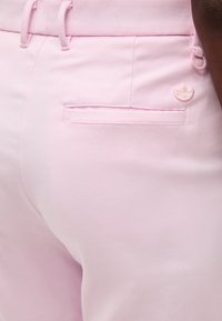 Ανοιχτό ροζ παντελόνι με πλαϊνή τσέπη στο πίσω μέρος, θηλιές για ζώνη και μικρό λογότυπο Adidas Trefoil στη δεξιά πλευρά κοντά στην τσέπη.