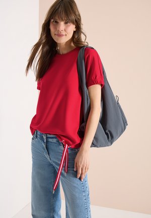 Młoda kobieta w czerwonej bluzce z krótkim rękawem i bocznymi troczkami, niebieskich dżinsach oraz szarej torbie na ramię na tle beżu i bieli.