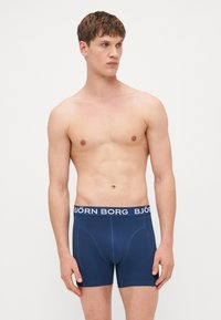 Temno modri bokserji z mehko teksturo, ki imajo bel "BJÖRN BORG" logotip na elastičnem pasu in prilegajočo se zasnovo.