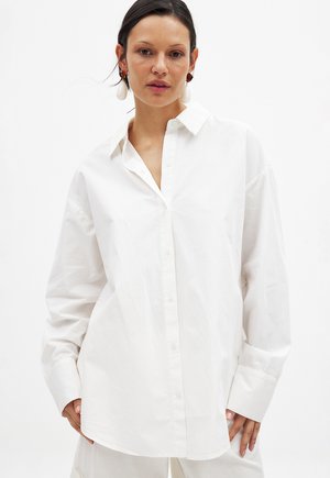 Biely, oversized button-up košeľa vyrobená z hladkej tkaniny, s klasickým golierom a dlhými rukávmi so širokými manžetami.