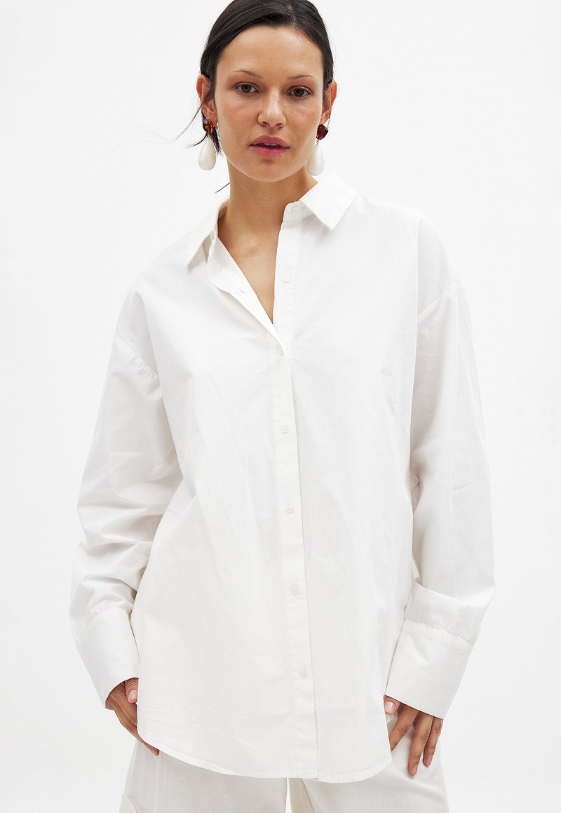 White, oversized button-up shirt made of smooth fabric, featuring a classic collar and long sleeves with wide cuffs.