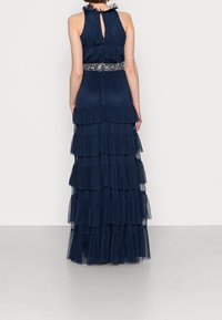 Mörkblå maxiklänning med lager av volanger, genomskinlig textur och detaljer med pärlor i midjan. Nyckelhålsrygg ger en unik designaspekt.
