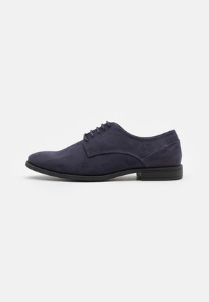 Chaussure Oxford pour homme en daim bleu marine avec semelle et lacets noirs, vue de côté sur fond blanc.