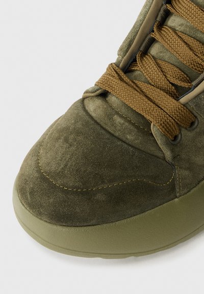 Olive green suede sneaker featuring a textured upper, raised stitching, and brown laces. Cushioned sole with a rounded toe design.