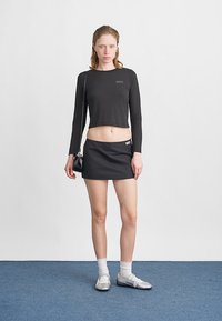 Junge Frau, die drinnen steht und ein schwarzes, kurzes Langarmoberteil, einen schwarzen Minirock, weiße Socken, silberne Schuhe trägt und eine kleine schwarze Tasche hält.