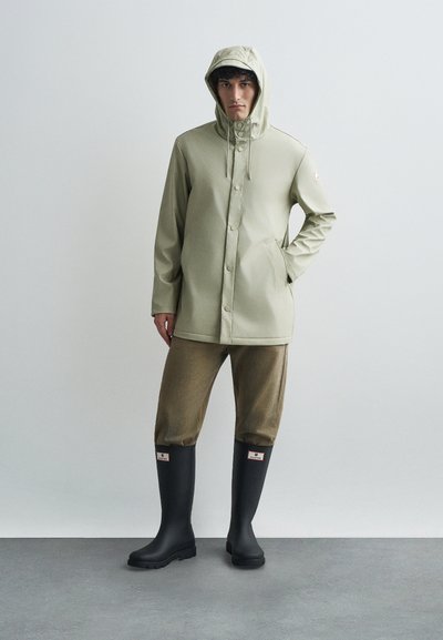 Svijetlozelena vodootporna jakna s kapuljačom, gumbima na dugme i bočnim džepovima, uparena s smeškim hlačama i crnim gumiranim čizmama.