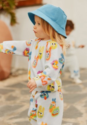 Fleece-Outfit mit bunten, cartoonartigen Tiergesichtern auf weißem Hintergrund. Das Kind trägt einen blauen Hut und hellgraue Hosen.