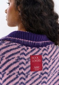 Kobieta o kręconych ciemnych włosach nosi sweter z fioletowo-różowym wzorem zygzaków z czerwoną metką z napisem „MIŁOŚNICY KSIĄŻEK”.