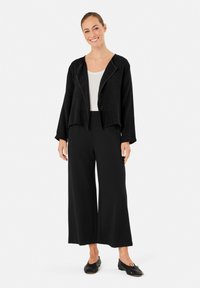 Zwarte cropped jack met een textuurafwerking, gecombineerd met wijde zwarte broek en zwarte flats. De top heeft een ronde halslijn en lange mouwen.