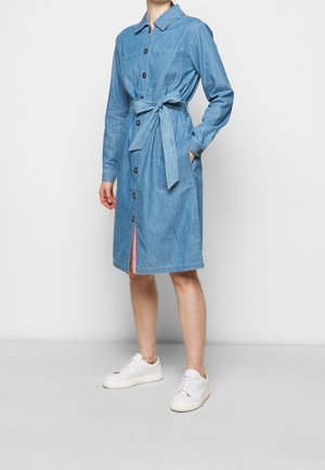 Woman wearing knee-length buttoned denim dress with long sleeves and tied waist belt, paired with white sneakers.