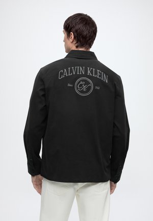 Mand iført en sort langærmet skjorte med teksten "Calvin Klein Since 1968" på ryggen og hvide bukser mod en ensfarvet baggrund.