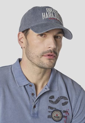 Hombre con barba ligera que lleva una camisa tipo polo azul y una gorra gris bordada con "HARAJUKU" y texto rojo sobre un fondo gris liso.