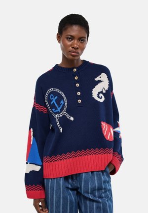 Femme portant un pull bleu marine avec une bordure rouge ornée de motifs nautiques incluant une ancre, un hippocampe, un coquillage et un voilier, associé à un pantalon rayé bleu.