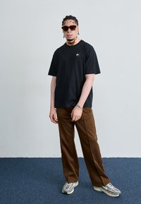 Black oversized t-shirt with a small logo, brown trousers, and gray sneakers. Accessories include dark sunglasses and a silver bracelet.