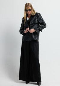 Donna che indossa pantaloni larghi neri, giacca in pelle nera, scarpe nere a punta e occhiali da sole cat-eye neri su uno sfondo chiaro e semplice.