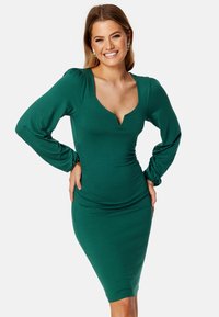 Zelené přiléhavé šaty vyrobené z elastické tkaniny, s dlouhými volánovými rukávy, hlubokým výstřihem a řasením po stranách.