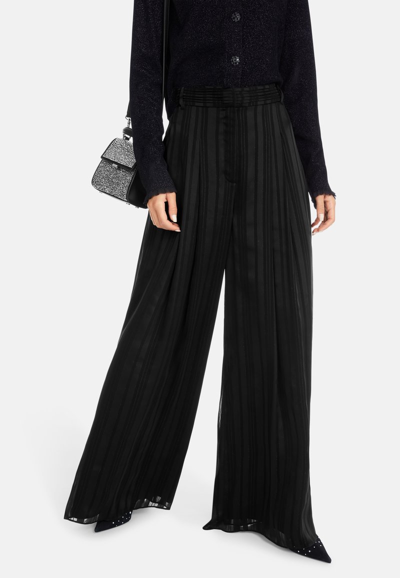 Schwarze weite Hose mit vertikalen Streifen, aus einem leichten Material. Kombiniert mit einem marineblauen strukturierten Pullover und einer kleinen Handtasche.