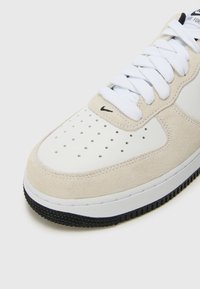 Beige suede and white leather sneaker with perforated toe, flat white laces, black swoosh, and textured black rubber sole.
