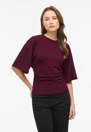Kobieta o długich brązowych włosach, ubraną w luźną bordową bluzkę z krótkim rękawem i czarne spodnie, stoi z jedną ręką za plecami.