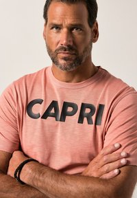 Lyserød kortærmet t-shirt med "CAPRI" i fede sorte bogstaver på forsiden. Stoffet ser blødt ud, med en rund halsudskæring og løstsiddende pasform.