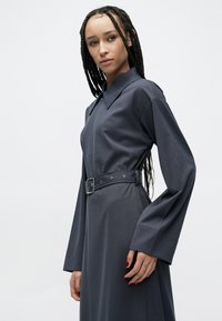 Tmavosivé šaty s dlhým rukávom, golierovým výstrihom, opaskom v páse a rozšírenými rukávmi. Hladká textúra s minimalistickým dizajnom.