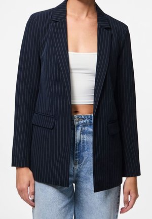 Frau trägt ein marineblaues Blazer mit Nadelstreifen über einem weißen, bauchfreien Top und hellblauen, hoch taillierten Jeans, abgebildet von Hals bis Hüfte.