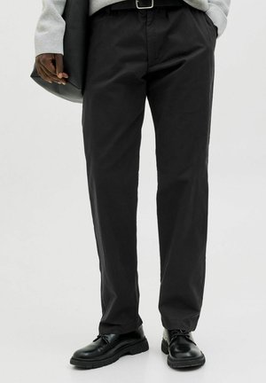 Black cotton trousers with a relaxed fit, featuring a waistband with belt loops and side pockets. Paired with black lace-up shoes.