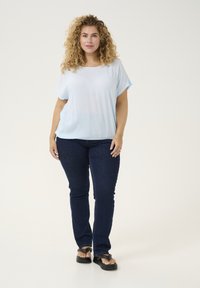 Top azzurro chiaro a maniche corte realizzato in tessuto morbido, abbinato a jeans skinny blu scuro e infradito nere, in piedi su uno sfondo semplice.