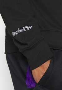 Manche de sweat-shirt noire avec broderie blanche "Mitchell & Ness" et main dans la poche d'un pantalon noir avec un détail de fermeture éclair violette.