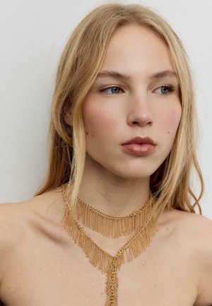 Mlada žena s dugom plavom kosom, noseći slojevite ogrlice od zlatnih lanaca s resama, gleda u stranu na jednostavnoj svijetloj pozadini.