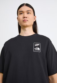 T-shirt en coton noir à manches courtes, avec un patch logo blanc sur la poitrine bordé d'un contour rectangulaire. Texture lisse, coupe décontractée.