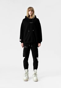 Sudadera negra de gran tamaño con un bolsillo canguro y cordones, combinada con pantalones de chándal negros que tienen acentos gráficos blancos y bolsillos con cremallera. Botas blancas.