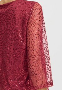 Przezroczysta czerwona bluzka z abstrakcyjnymi wzorami z cekinów, posiadająca półprzezroczyste długie rękawy i dopasowany dekolt.