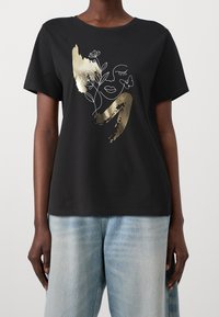 En person som bär en svart t-shirt med en guld- och vit abstrakt ansiktsdesign samt blommor och fjärilar, tillsammans med ljusblå jeans.