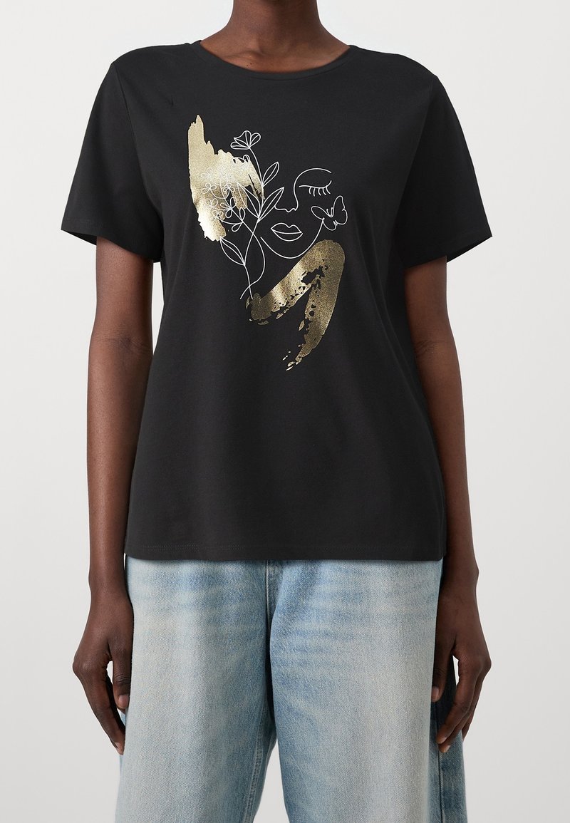 En person som bär en svart t-shirt med en guld- och vit abstrakt ansiktsdesign samt blommor och fjärilar, tillsammans med ljusblå jeans.