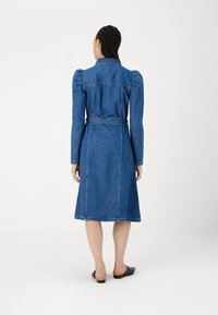 Vestido de ganga com mangas longas, design de ombros baloné, cintura cingida e saia até ao joelho. Tecido azul com detalhes de costura visíveis.