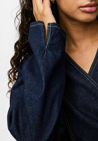 Top in denim blu scuro con scollatura a V e maniche lunghe, caratterizzato da cuciture dorate a vista e una finitura leggermente testurizzata.