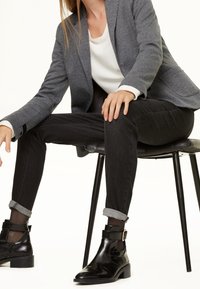 Harmaa teksturoitu bleiseri, valkoinen yläosa, mustat kapenevat farkut käänteillä ja kiiltävät mustat nilkkurit solkikoristeella, istuu mustalla tuolilla.