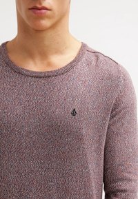 Trui in een gestructureerde brei met een bordeauxrode en grijze kleurmix. Heeft een ronde halslijn en een klein zwart logo op de borst.