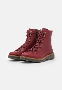Botas de tobillo de cuero rojo con suela de goma texturizada, que cuentan con un diseño de cordones en la parte frontal, ojetes metálicos y una cremallera lateral para facilitar su uso.